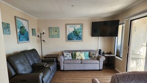 Una sala de estar con un sofá gris, un sofá de cuero negro y un sillón gris. Hay un televisor de pantalla plana montado en la pared, una lámpara de pie y obras de arte en las paredes.