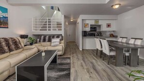 Una sala moderna con un sofá, una mesa de centro y un comedor con una mesa y sillas. Una escalera conduce a un nivel superior.