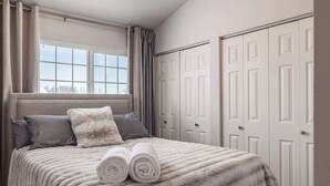 Ein Schlafzimmer mit einem Bett, zwei Kissen, einem zusammengerollten Handtuch, einem Fenster und einer Reihe weißer Türen.