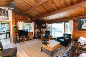 Ein gemütliches Blockhaus-Interieur mit Holzbalkendecke, Kamin, Fernseher und einer komfortablen Sitzecke mit Sofa und Sesseln.