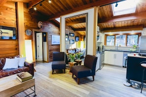 Ein gemütliches Blockhaus-Interieur mit Holzbalkendecke, einem Wohnbereich mit Ledersofa, zwei Sesseln, einem Couchtisch und einer Küche mit großem Fenster.