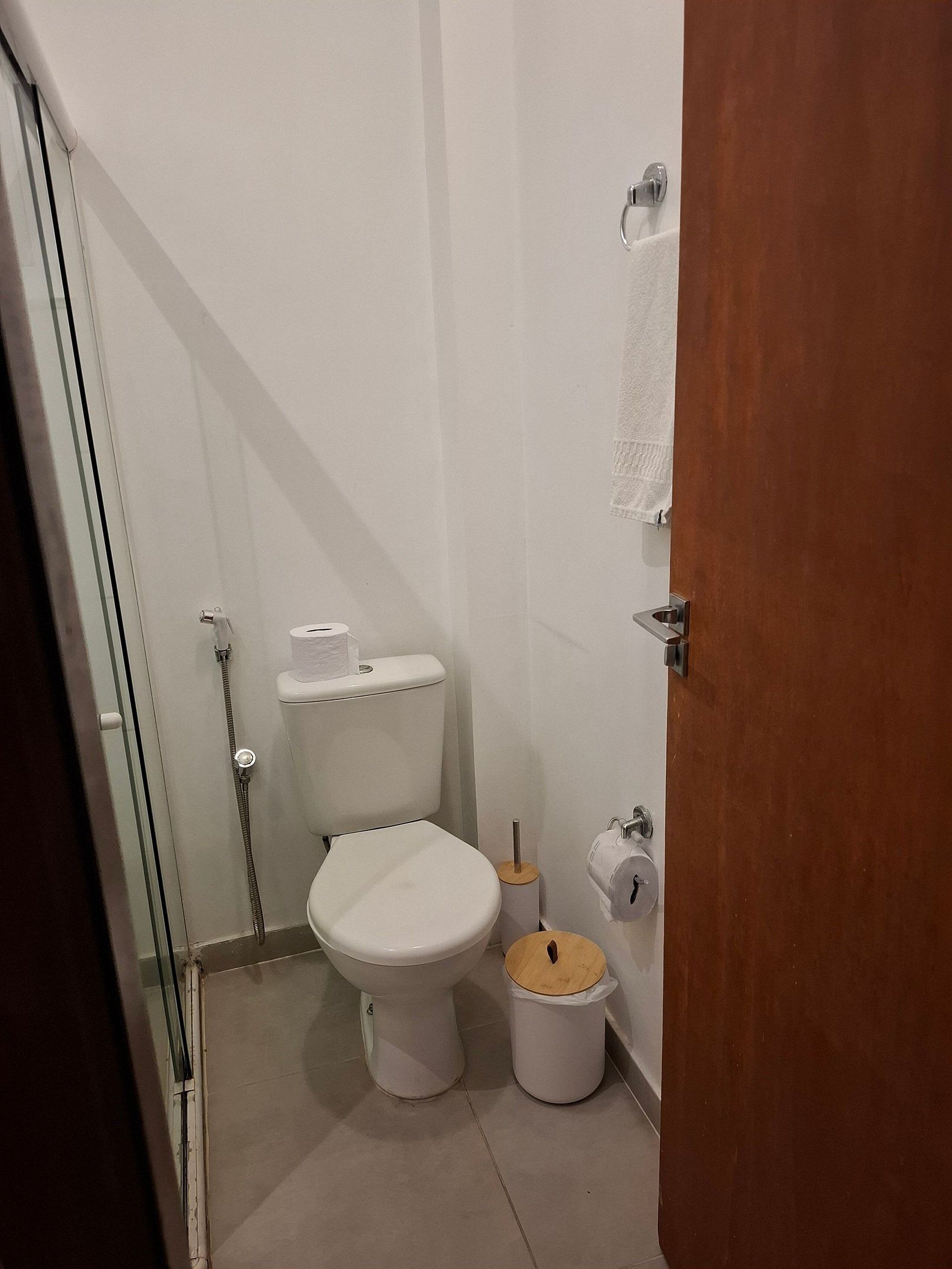 Un baño pequeño con inodoro, ducha, perchero y un cubo de basura.