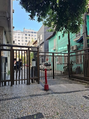Uma entrada protegida por portão e cerca de metal, calçada de paralelepípedos e edifícios ao fundo.