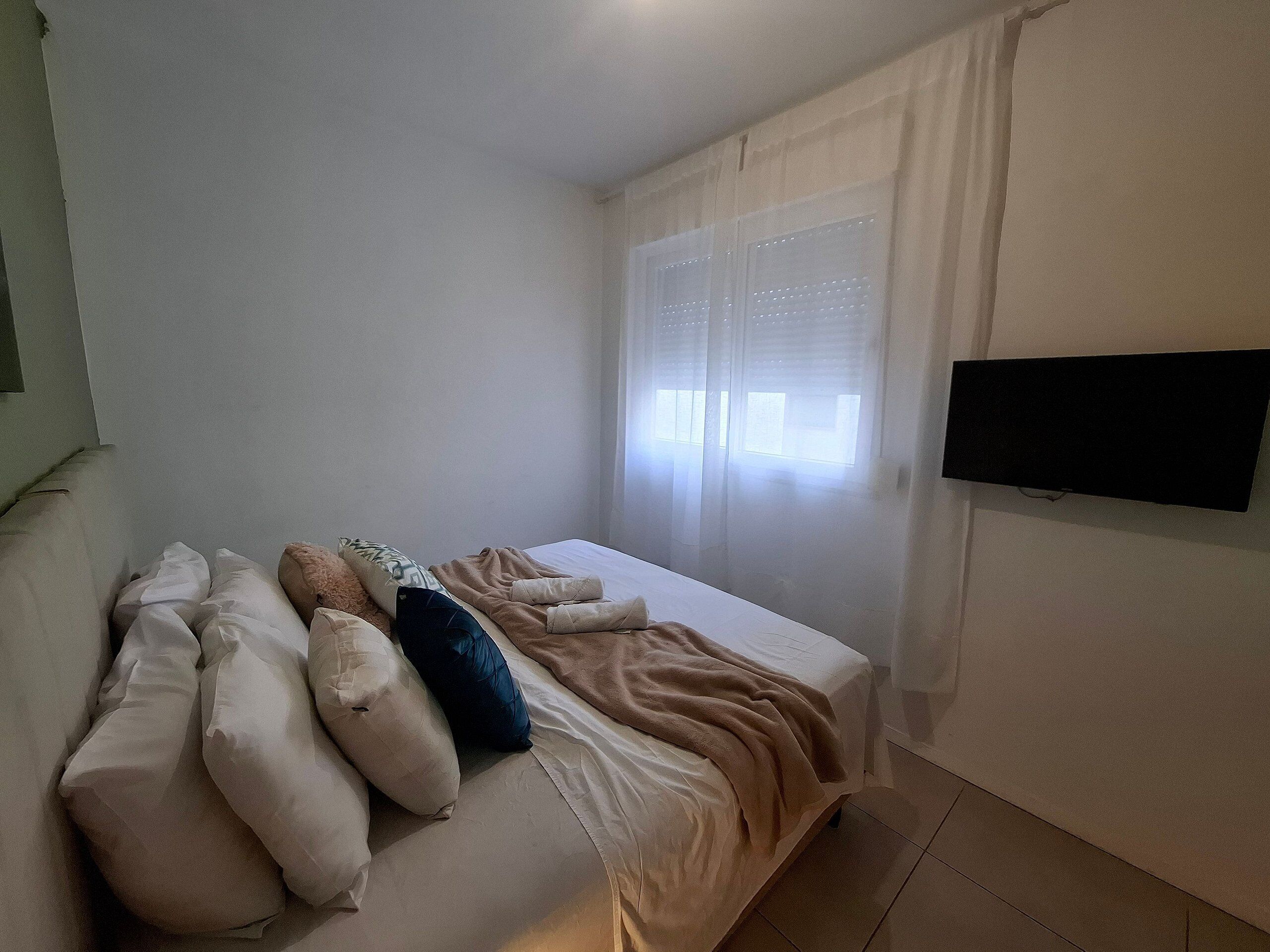 Un dormitorio con cama, almohadas, manta, televisor montado en la pared y ventana con cortinas.