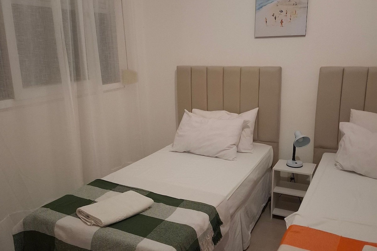 Habitación de hotel con dos camas, una mesita de noche con lámpara, un cuadro en la pared y una ventana con cortinas translúcidas.