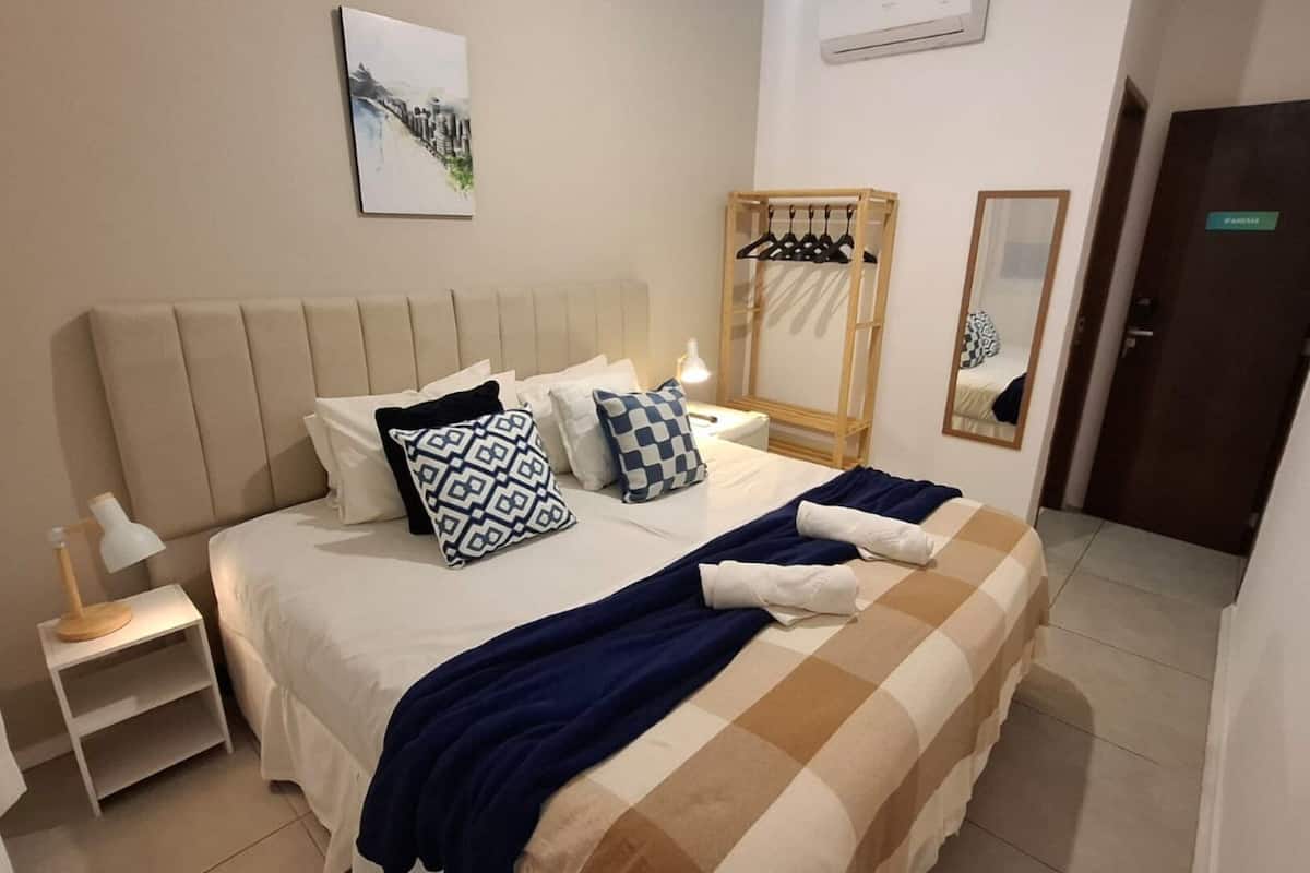 Una habitación de hotel con cama, mesita de noche, lámpara, obra de arte, espejo y armario.