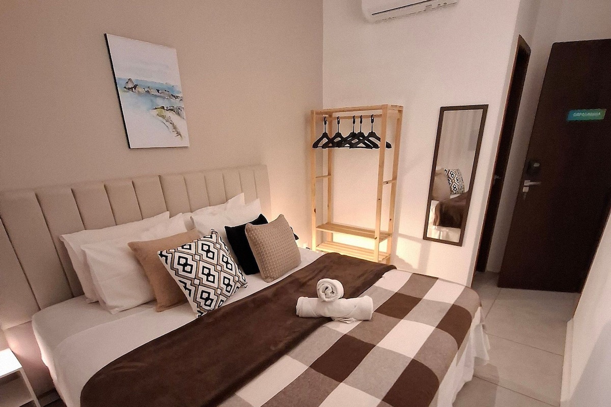 Una cama bien hecha con colcha a cuadros, un perchero, una mesita de noche y un aire acondicionado instalado en la pared.