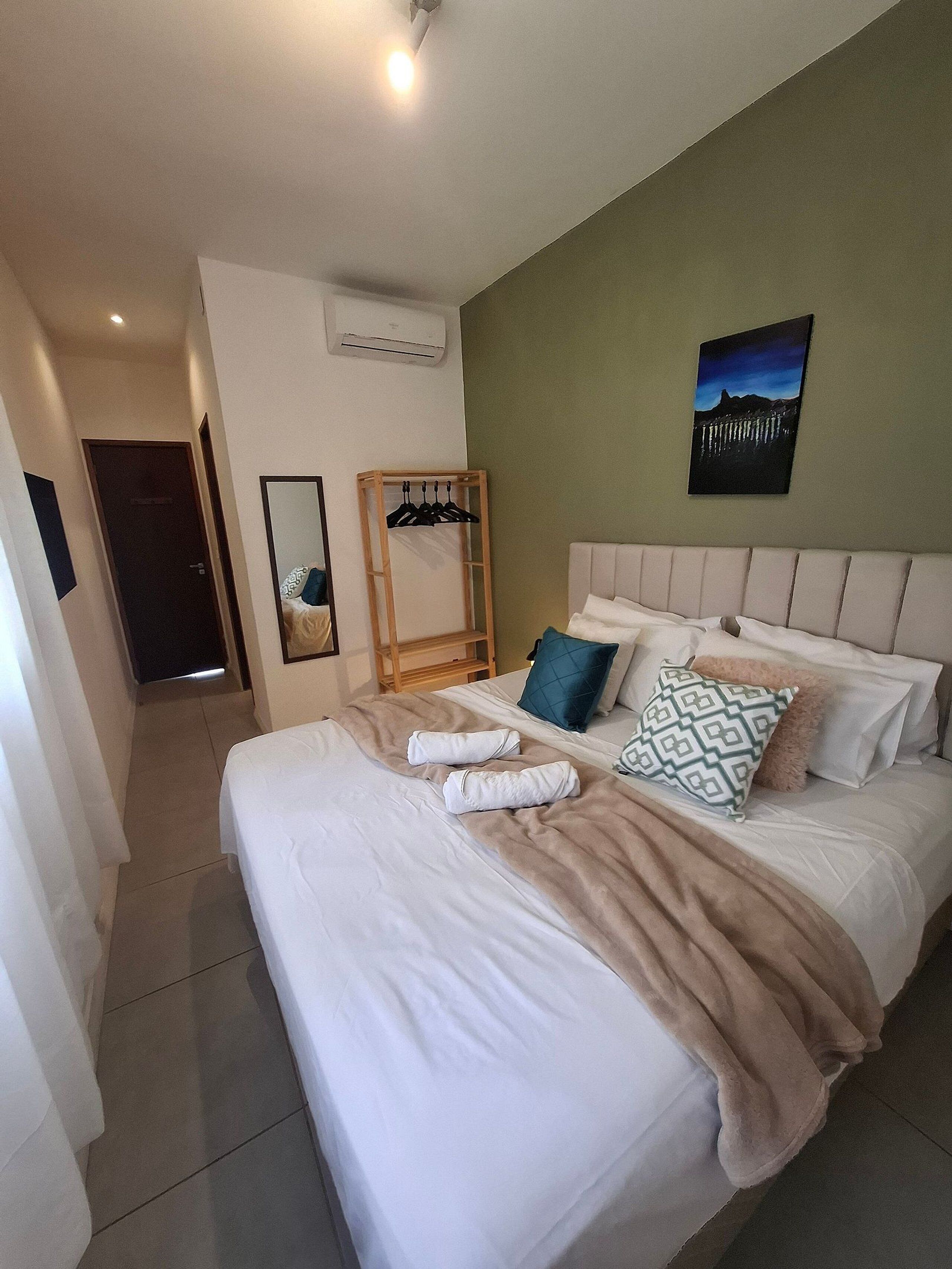 A hotel room with a large bed, a wooden hanger rack, a painting, and an air conditioning unit.