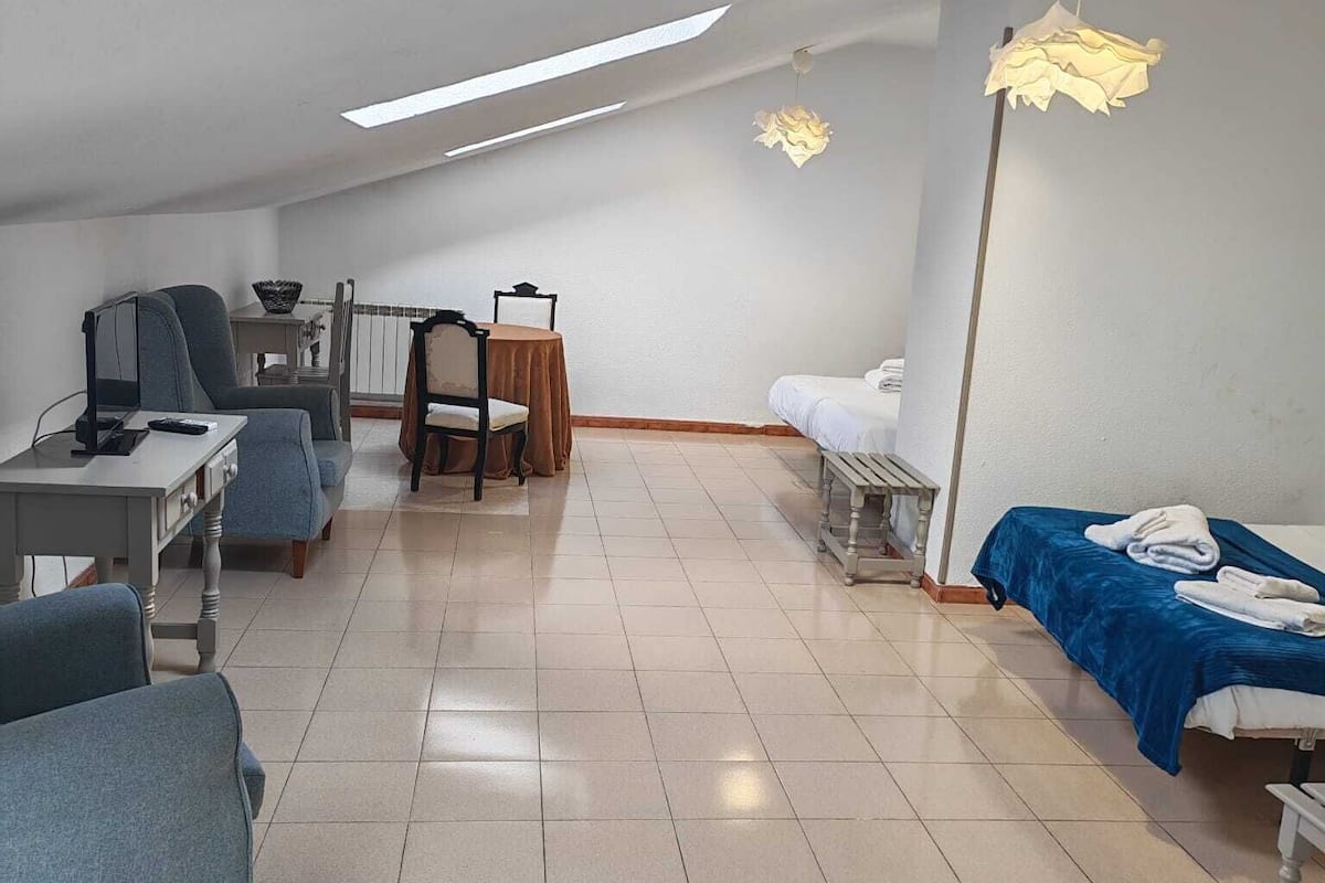 Ein geräumiges Zimmer mit hoher Decke, einer Essecke mit Tisch und Stühlen, einem Bett mit blauer Decke sowie einem Schreibtisch mit Stuhl.