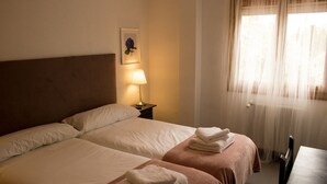 Une chambre d’hôtel avec deux lits, une table de chevet avec une lampe, un tableau encadré au mur et une fenêtre avec des rideaux légers.