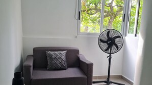 Um quarto com um sofá cinza, um ventilador de chão preto e uma pequena mesa com um vaso decorativo.