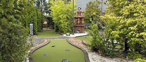 Un campo de mini golf con un sendero serpenteante, césped artificial verde y elementos decorativos como una estatua y una fuente.