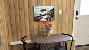 Ein Essbereich mit einer Holzwand, einem Gemälde einer Strandansicht, einem kleinen Tisch mit zwei Stühlen und einer Topfpflanze.