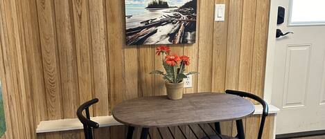 A dining area with a wooden wall, a painting of a beach scene, a small table with two chairs, and a potted plant.