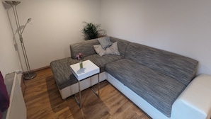 Een moderne woonkamer met een grijze modulaire bank, een klein wit bijzettafeltje, een vloerlamp en een potplant.