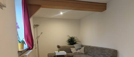 Een gezellige woonkamer met een grijze bank, een klein wit bijzettafeltje, een houten stoel en een raam met een rood gordijn.