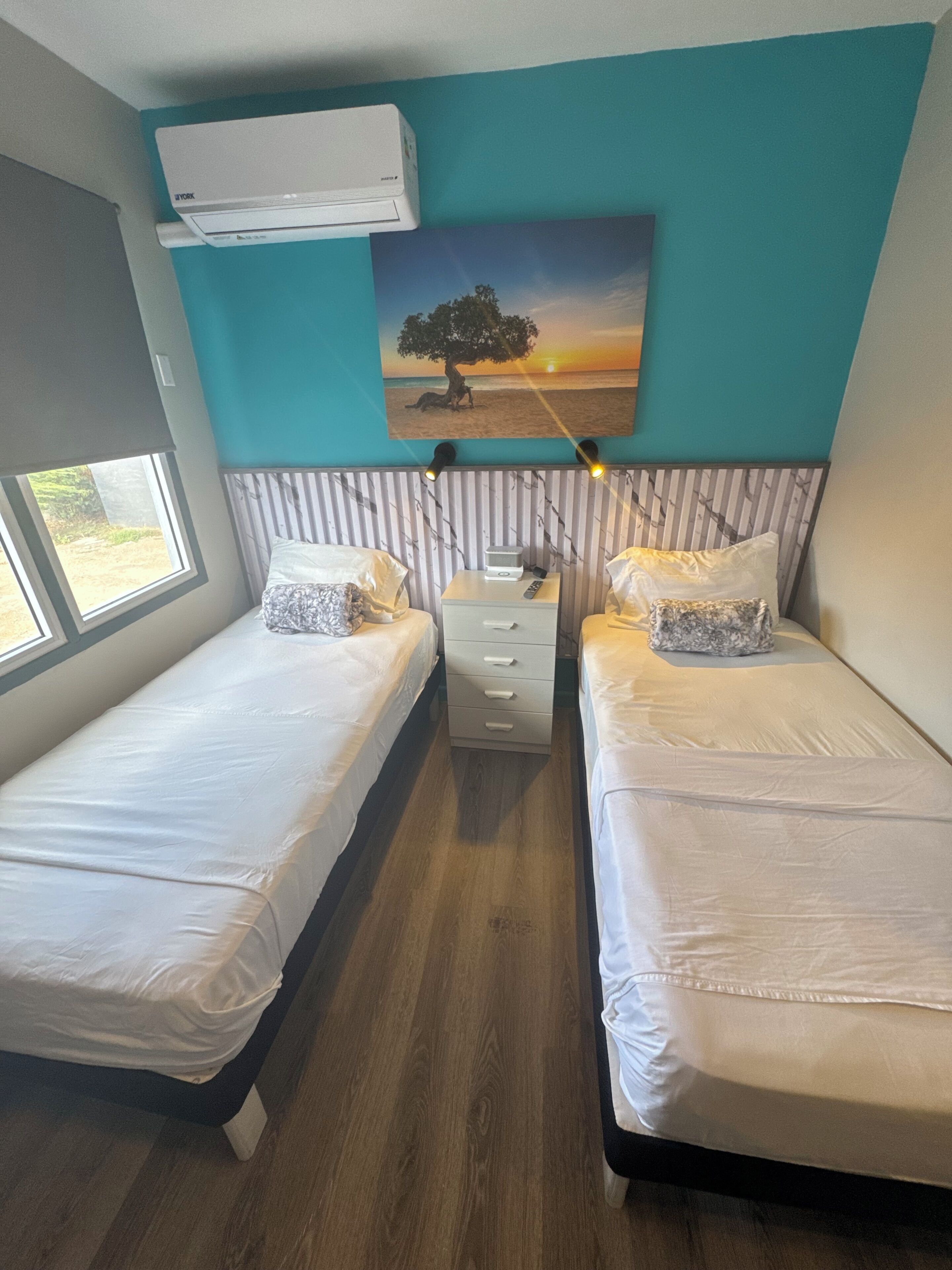 A bedroom with two single beds, a nightstand, an air conditioning unit, a painting of a tree by a beach, and a window with a view.