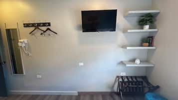 Habitación de hotel con una cama, un televisor montado en la pared, un perchero y una pequeña estantería con una planta y libros.