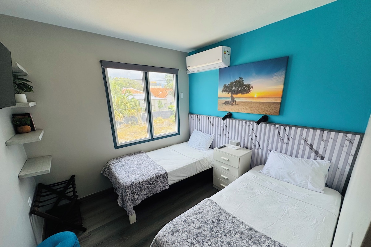 Un dormitorio con dos camas, una ventana, aire acondicionado instalado en la pared, una mesita de noche con lámpara y un cuadro de un árbol.