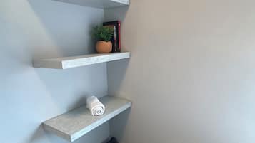 Rincón con pared blanca, un taburete azul, una silla de madera y un estante con una planta y un libro.