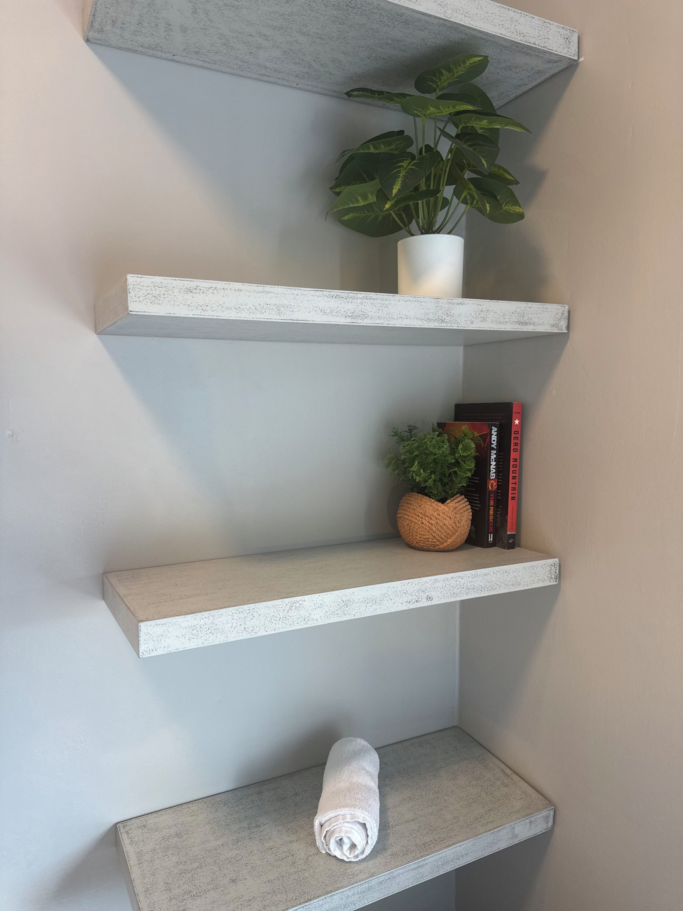 Eine Ecke mit drei weißen Regalen, auf jedem ein Topf mit einer Pflanze, ein Buch und ein zusammenrolltes Handtuch.