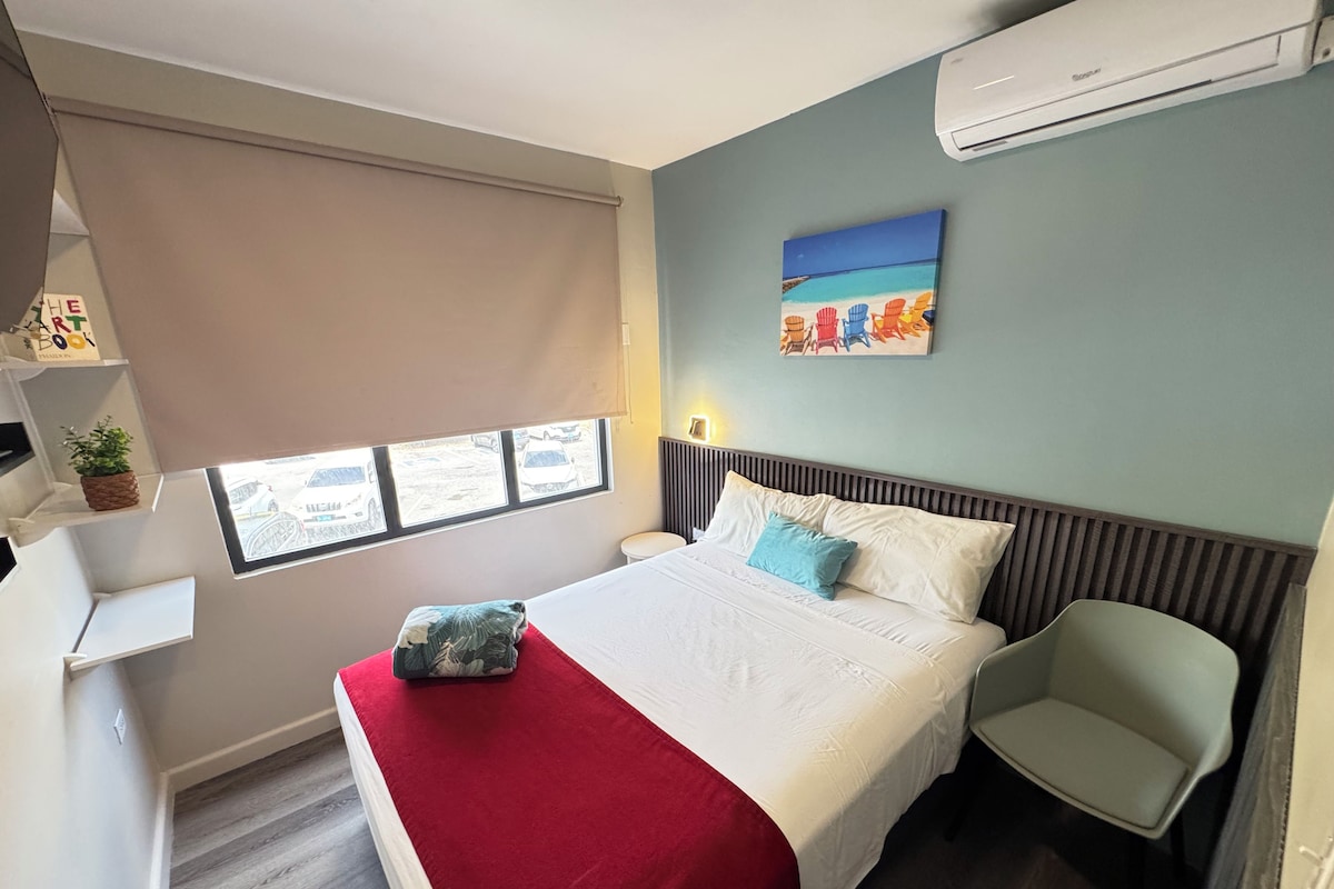 A hotel room with a bed, a chair, a window with a view, a painting on the wall, and a shelf with items.