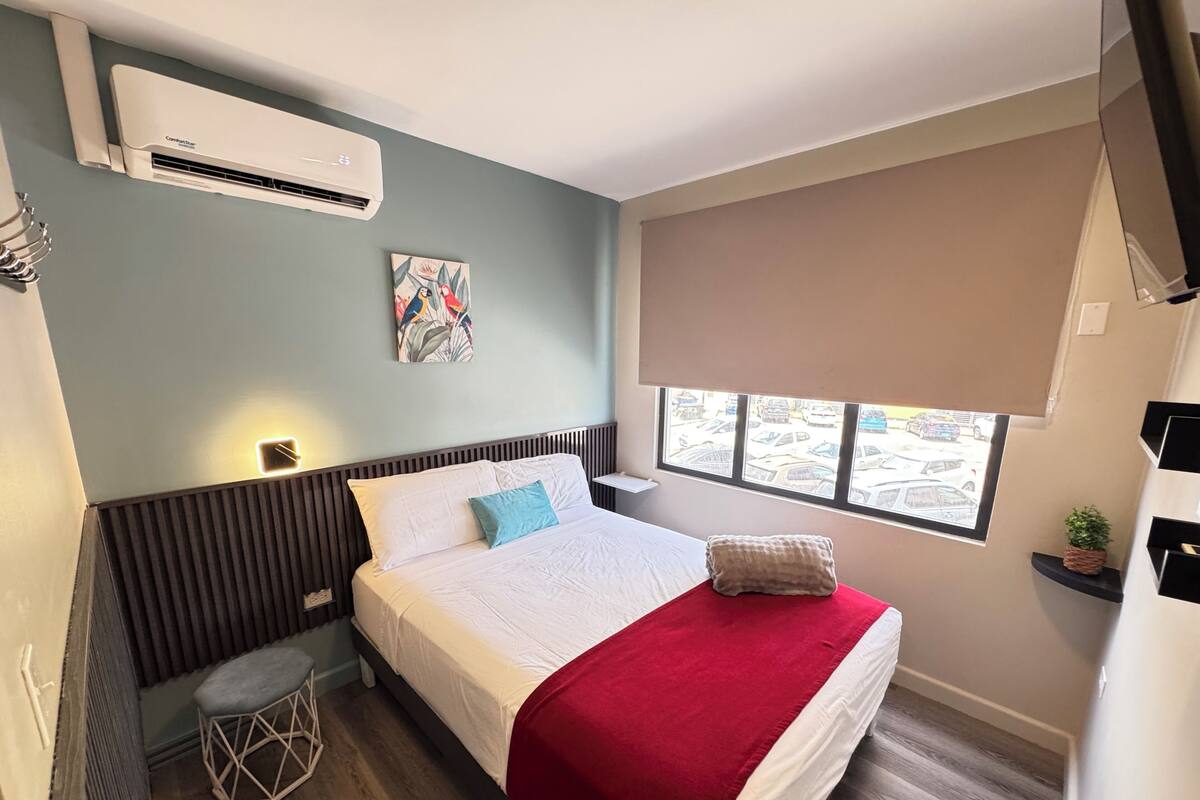 A hotel room with a bed, a window with a view of parked cars, an air conditioning unit, and a small stool.