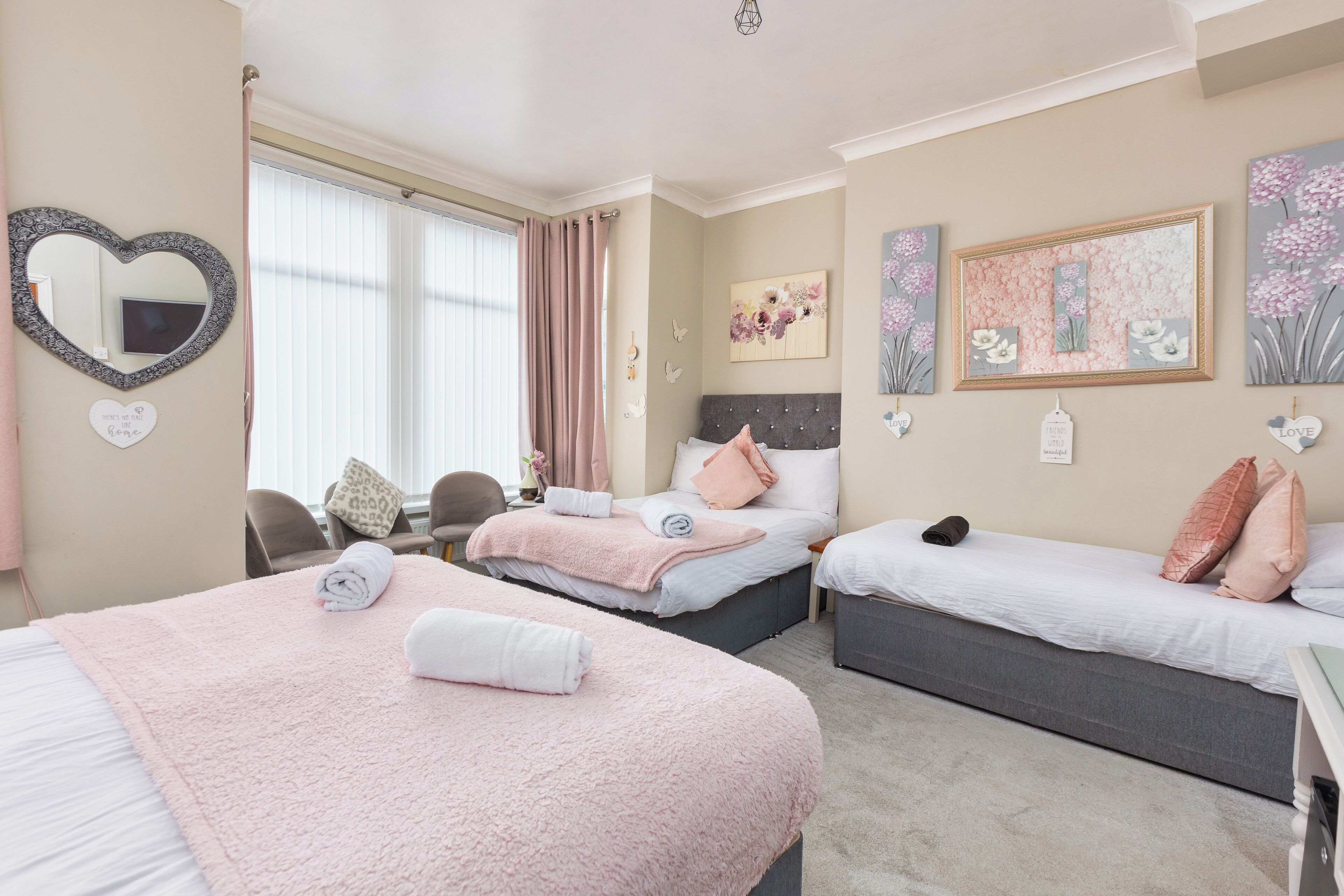 Habitación de hotel con dos camas, un espejo con forma de corazón, obra de arte floral y una pequeña mesa con sillas.