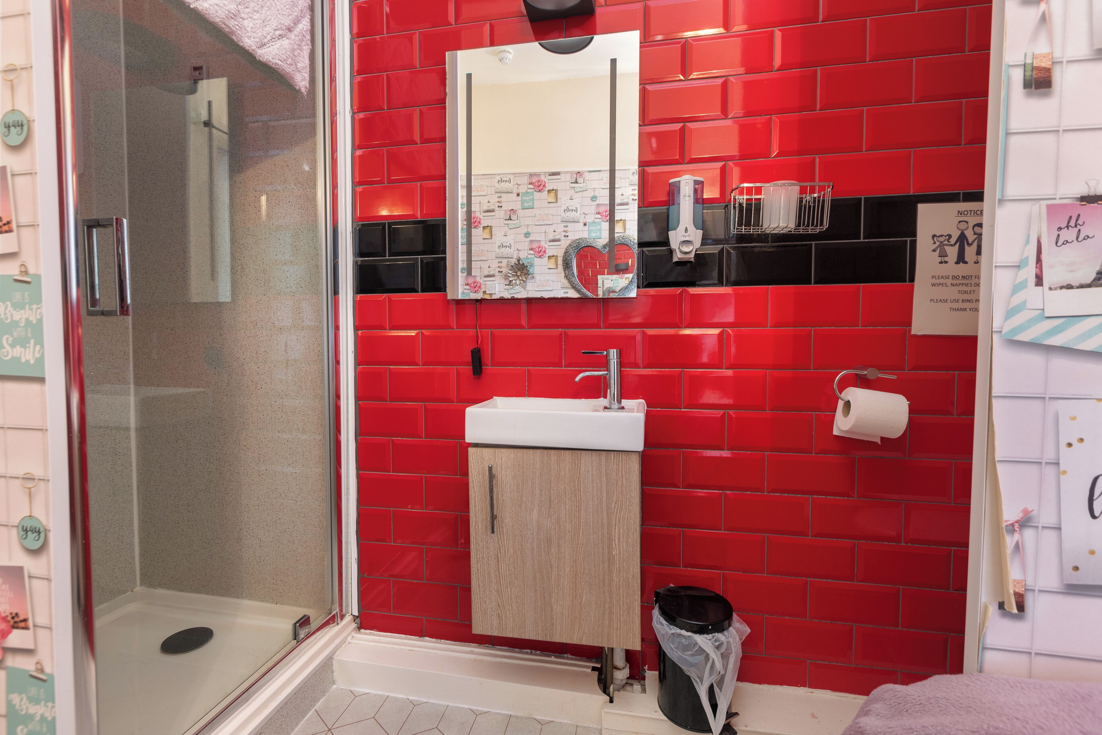 A bathroom with a red tiled wall, a glass shower enclosure, a sink with a mirror, and a toilet paper holder.