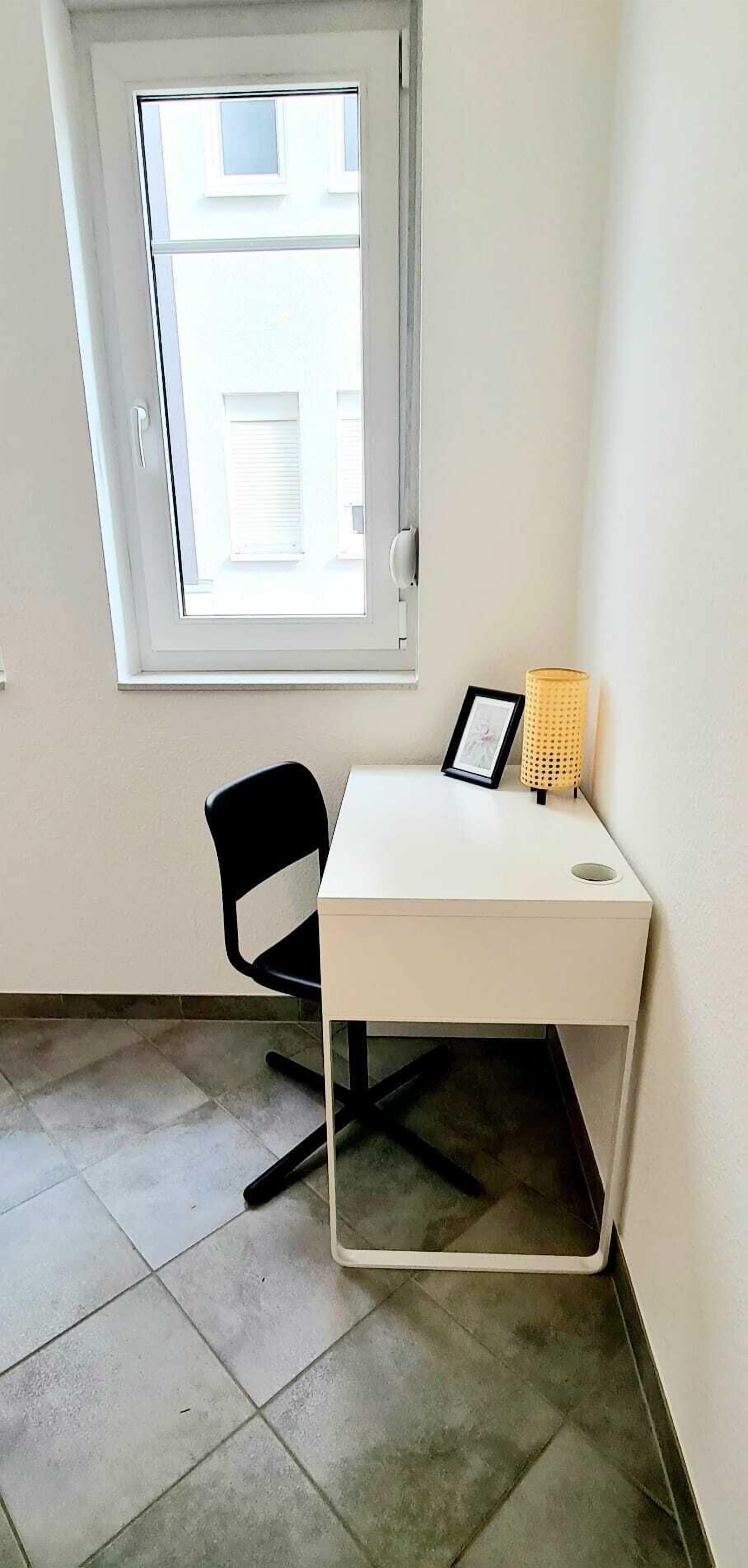 Una habitación pequeña con un escritorio blanco, una silla negra, una ventana y un marco de fotos sobre el escritorio.