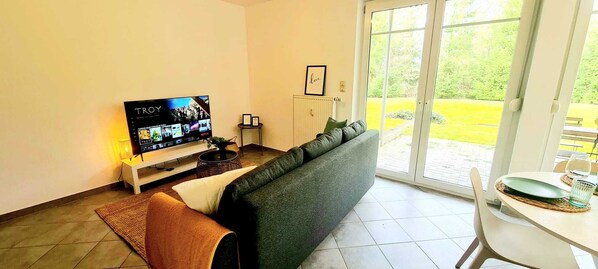 Ein Wohnzimmer mit Fernseher auf einem Schrank, einer Couch, einem kleinen Tisch mit Glas und Teller und Blick auf einen Garten durch eine Glastür.