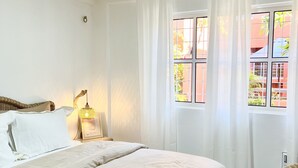 A bedroom with a large bed, a bedside lamp, a wicker chair, and a window with curtains. - Stay at Casa Carnaval walking distance to Playa Moreno (Pampatar)