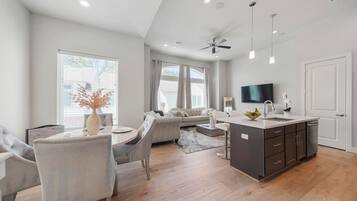 Een moderne keuken met een centraal eiland, een eetgedeelte met een ronde tafel en stoelen, en een zithoek met een bank en een aan de muur bevestigde televisie.