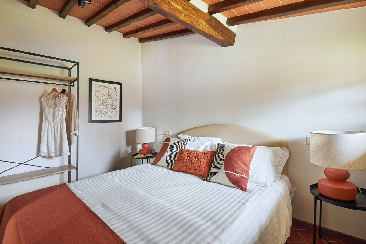 A neatly made bed with striped bedding, a red throw blanket, and multiple pillows, including a football-themed one. There is a wall-mounted rack with clothes, a framed abstract artwork, and a small table with a lamp. The room features exposed wooden beams on the ceiling.