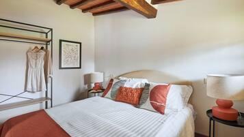 Una cama bien hecha con ropa de cama a rayas, un plaid rojo y varios almohadones, incluyendo uno con tema de fútbol. Hay un perchero de pared con ropa, una obra de arte abstracta enmarcada y una mesita de noche con una lámpara. La habitación cuenta con vigas de madera a la vista en el techo.