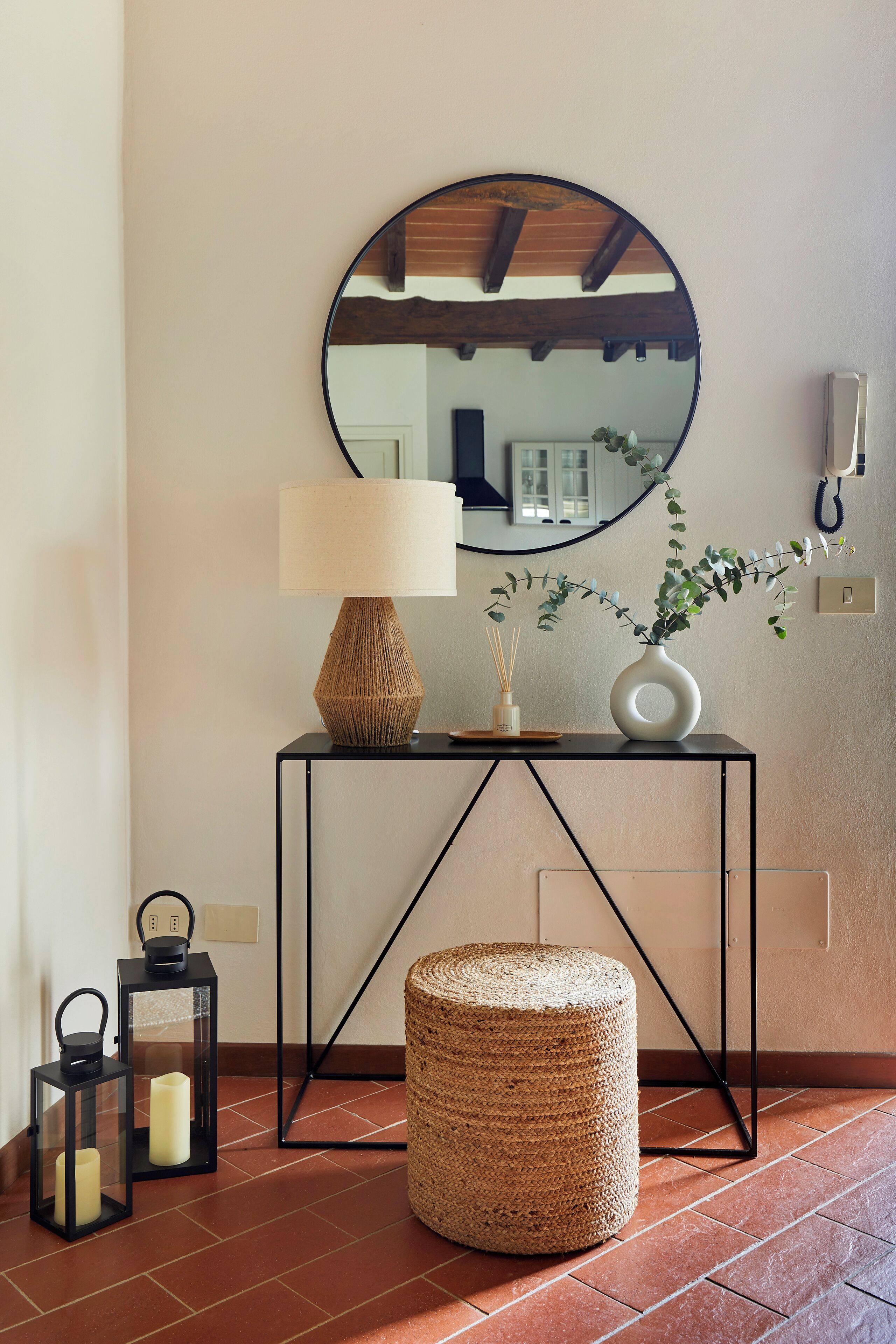 A modern interior with a round mirror, a lamp, a vase with a plant, and a small stool on a console table.