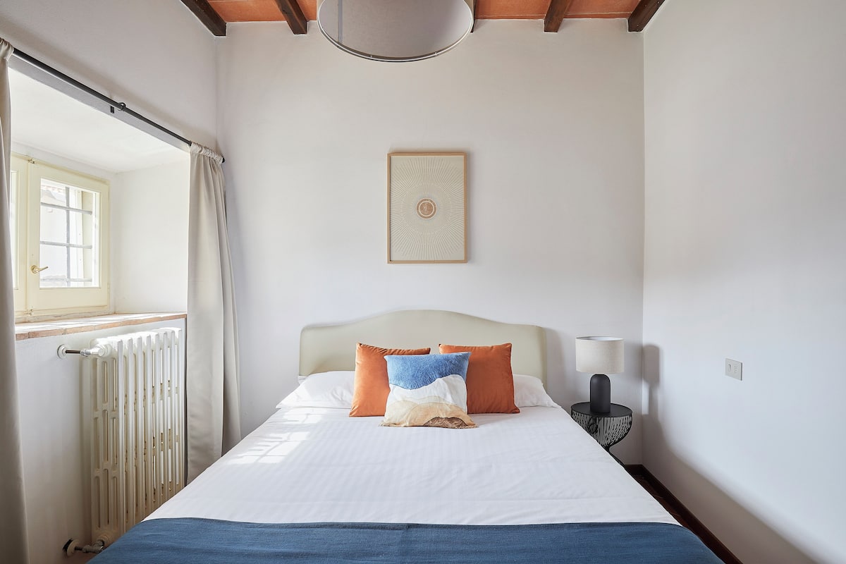 A neatly made bed with a white and blue pillow, an orange pillow, and a decorative piece above it.