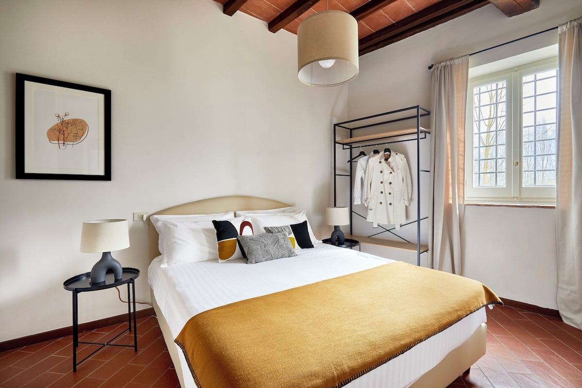 A neatly made bed with a white headboard, a gold throw blanket, and a variety of pillows. There is a framed artwork on the wall, a bedside table with a lamp, and a clothing rack with hanging garments.