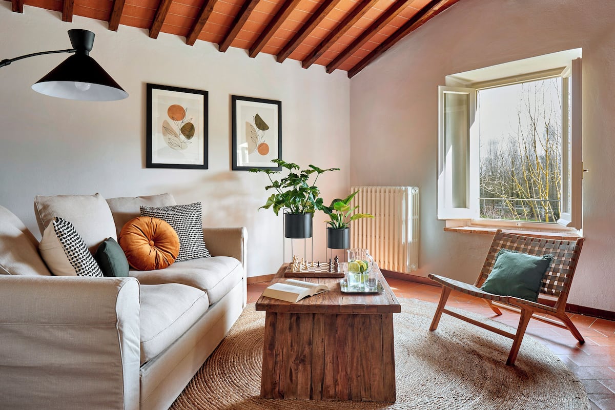 A cozy living room with a sofa, a wooden coffee table, a wicker chair, and a window with a view of trees.