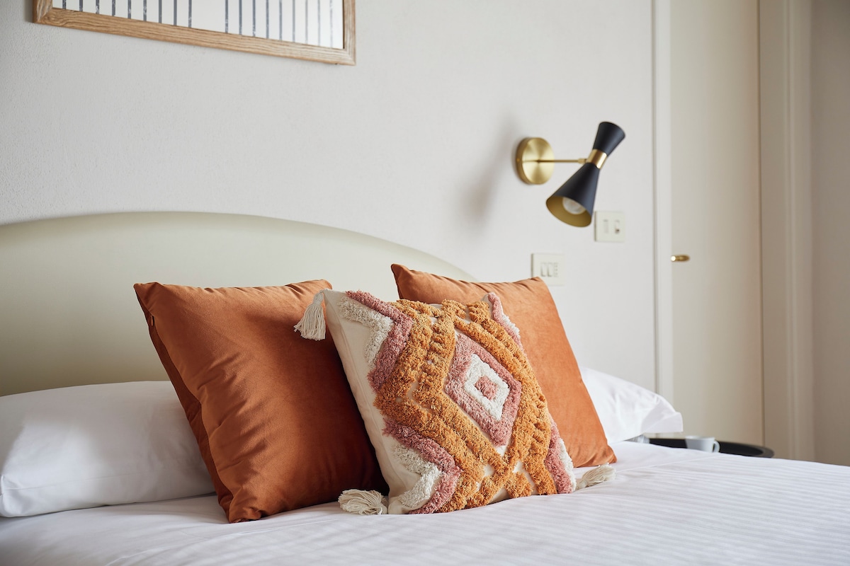 Una cama bien tendida con dos almohadas, una lámpara de pared y un cuadro enmarcado encima de la cama.