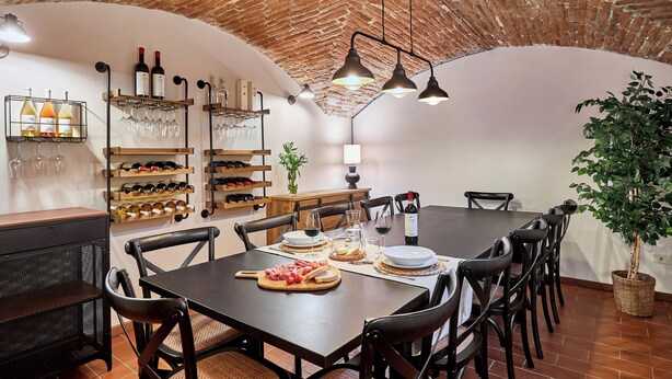 Un área de comedor con techo de ladrillo, mesas y sillas de madera. Se observan botellas de vino y copas en estanterías.