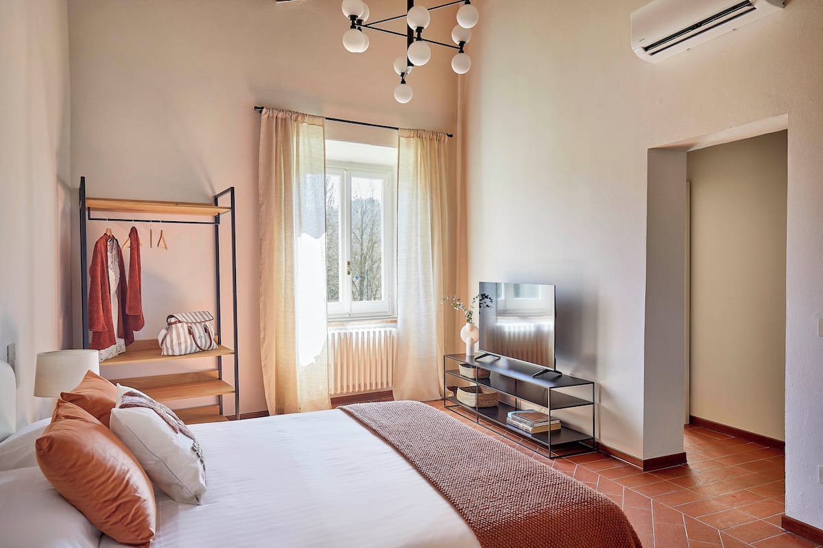A hotel room with a bed, a TV, a wardrobe, a window with curtains, and a radiator.