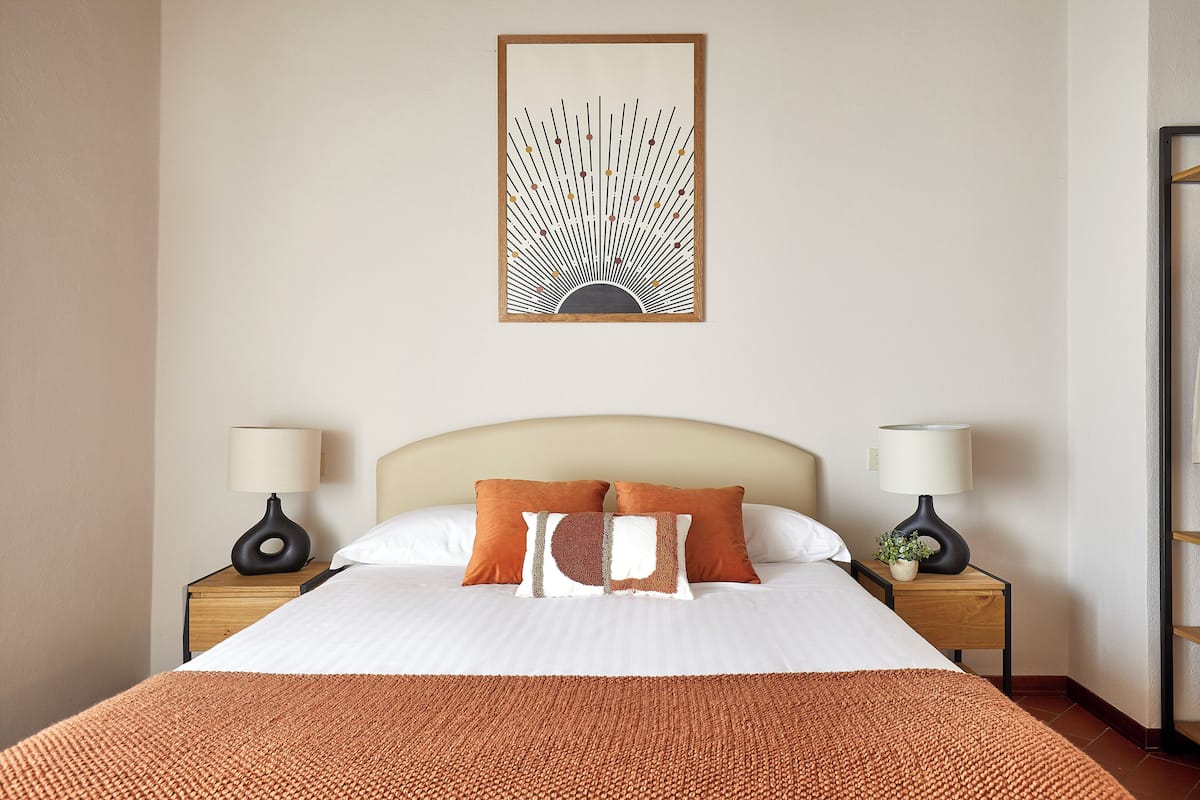 A neatly made bed with two pillows, a bedside table with a lamp, and a wall-mounted artwork.