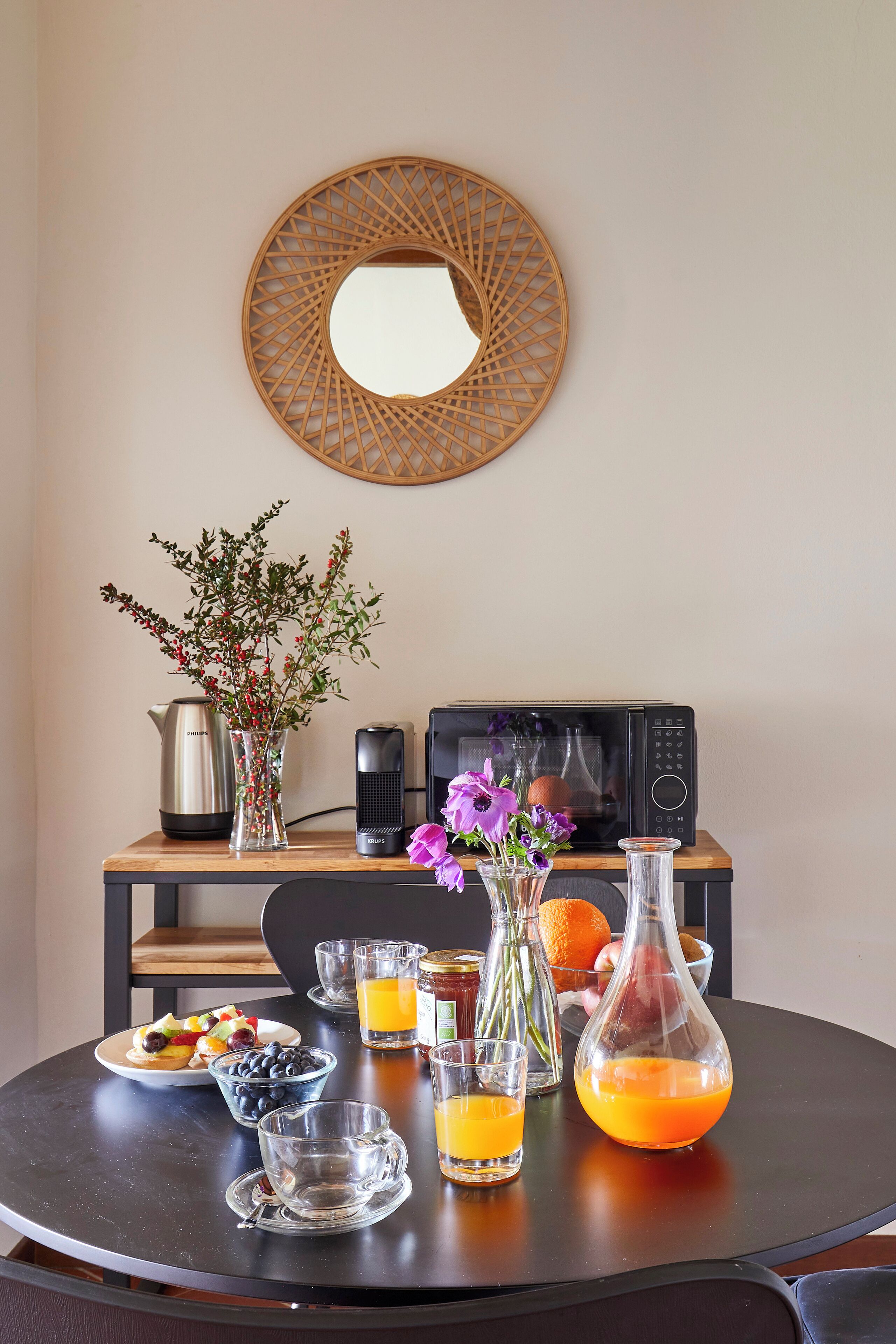 Una mesa de desayuno con una tetera, un microondas, un florero con flores, un cuenco de frutas y vasos de zumo de naranja.
