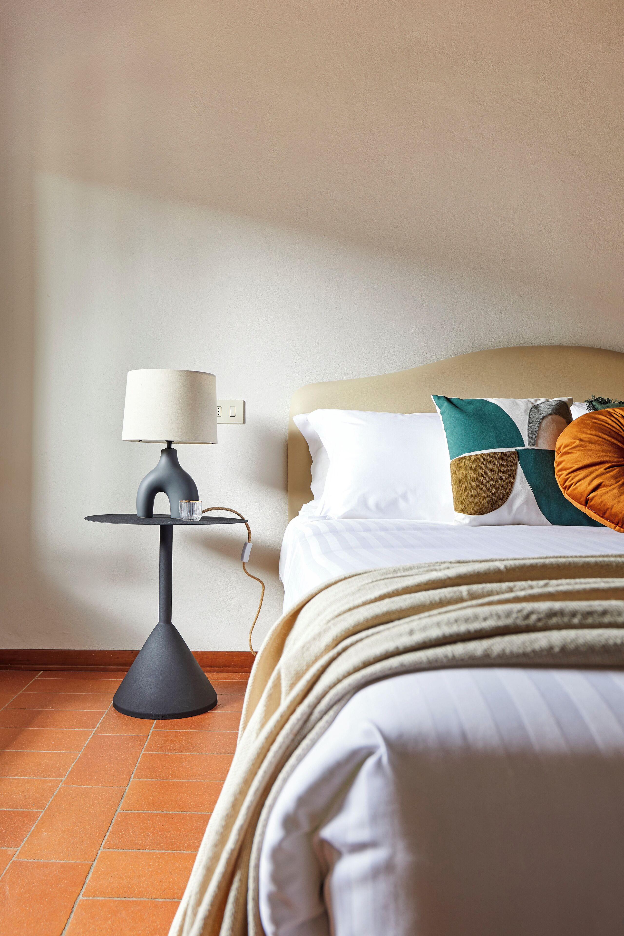 A neatly made bed with a white comforter and multiple pillows, a modern table lamp with a beige shade, and a tiled floor.