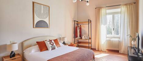 Un dormitorio con cama, mesitas de noche, lámpara colgante, perchero y una ventana con cortinas.