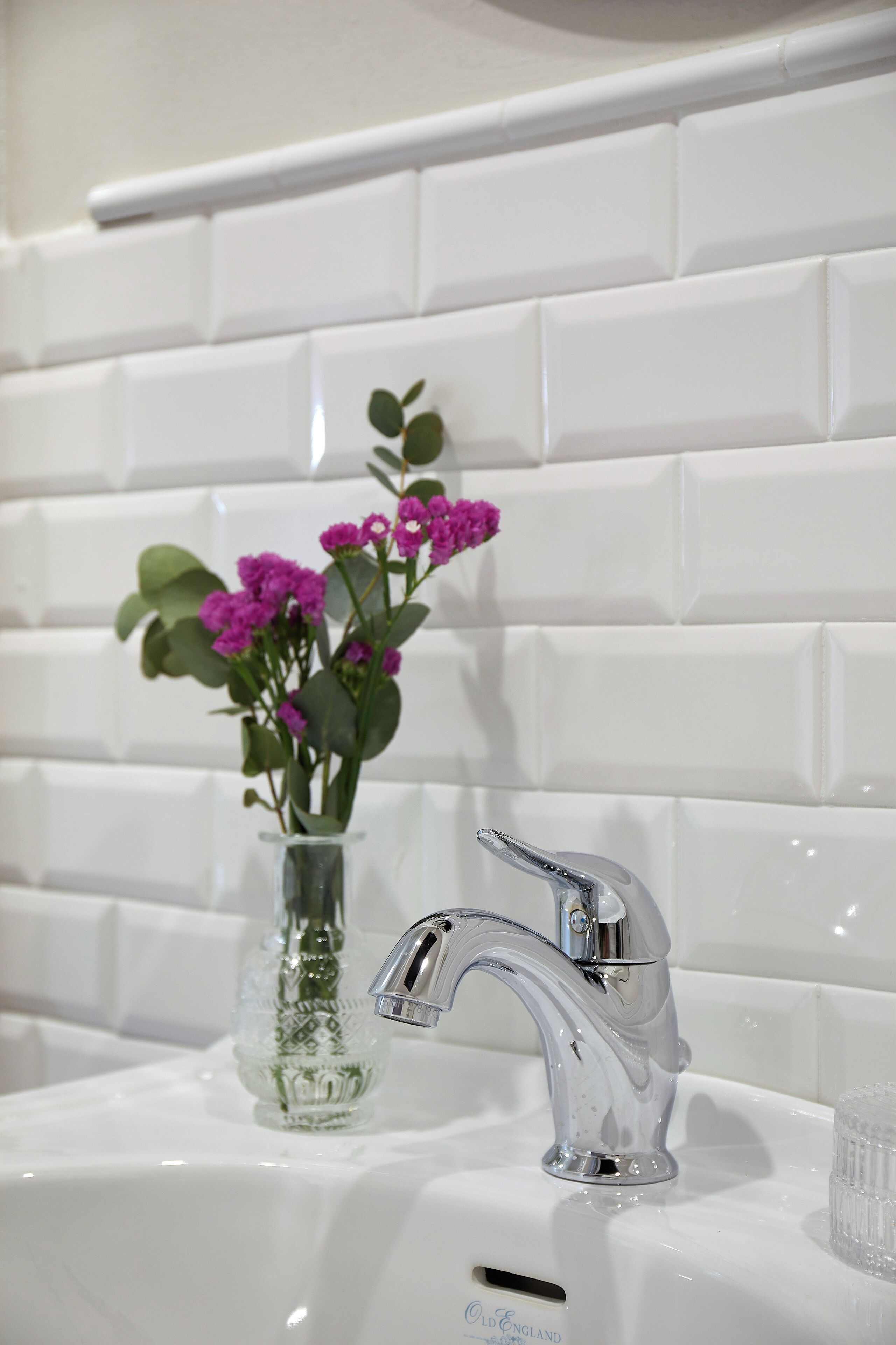 A bathroom with white tiled walls, a clear glass vase with purple flowers, and a modern chrome faucet.