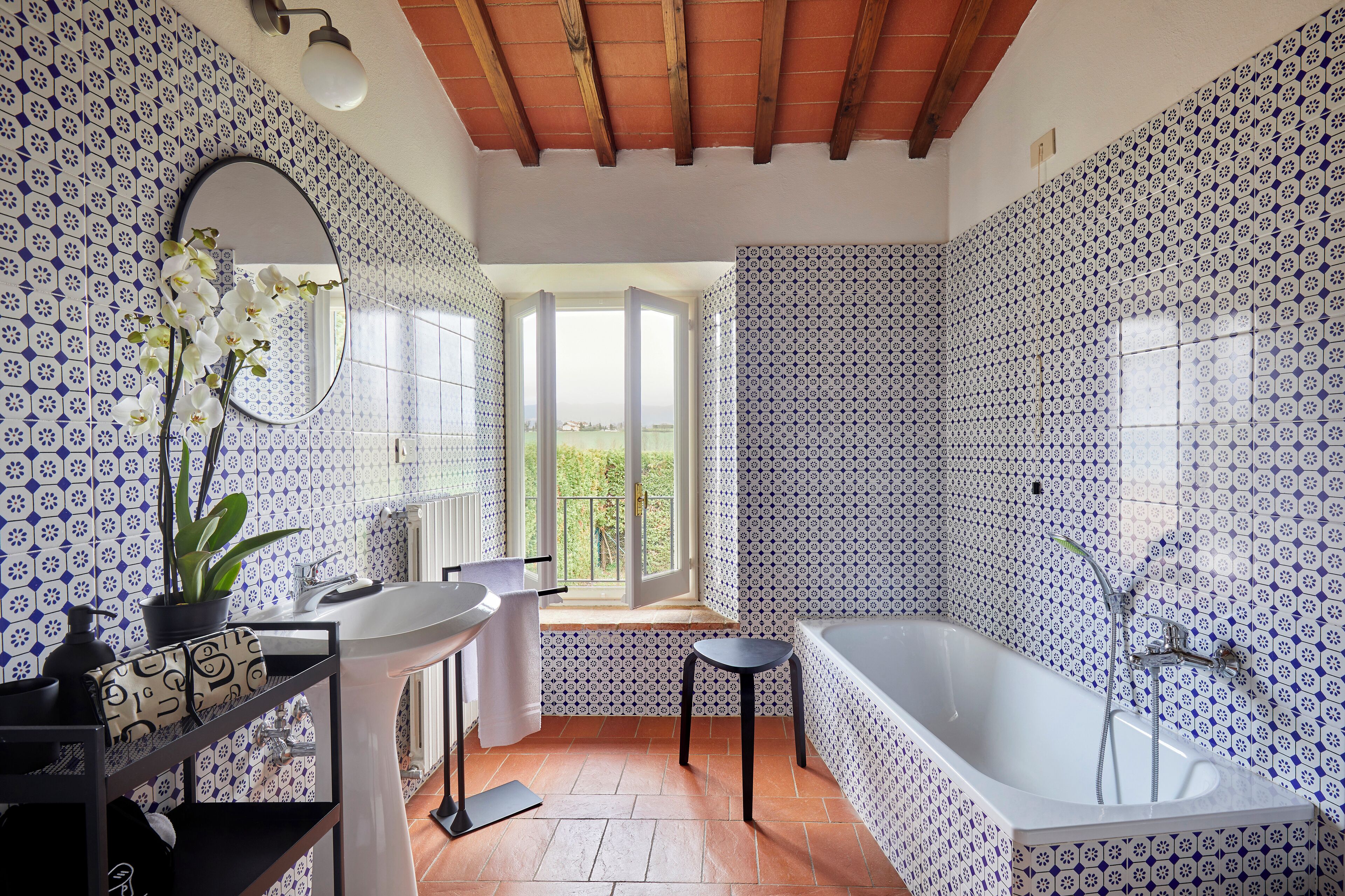 A bathroom with patterned tiles, a sink, a mirror, a potted orchid, a bathtub, and a window with a view.
