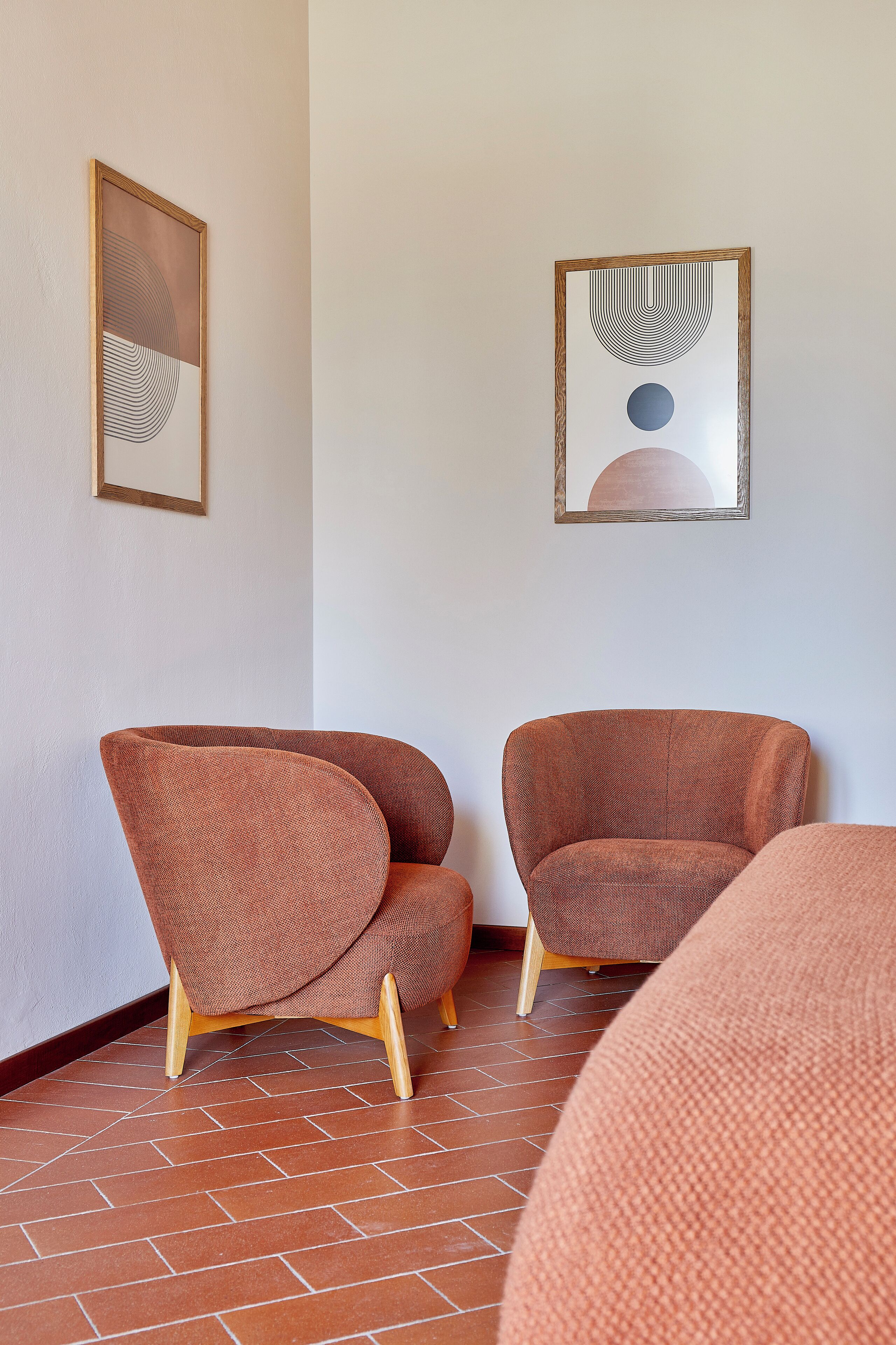Een kamer met twee bruine fauteuils op pootjes, een betegelde vloer en twee ingelijste kunstwerken aan de muur.