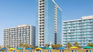 Un hotel con varios edificios altos, con balcones y una zona de playa con tumbonas y sombrillas.